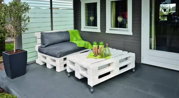 Sofá y mesa de centro hechos con palets pintados de blanco, con cojines grises y manta verde.