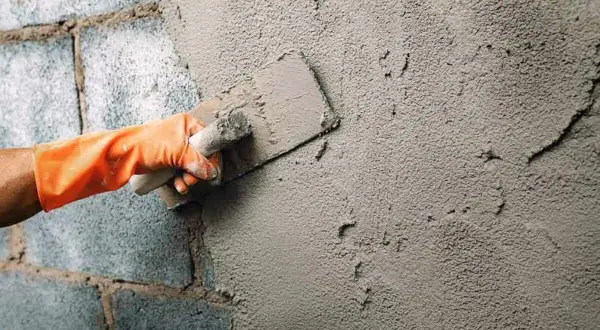 Mano con guante naranja aplica cemento para enfoscar pared exterior con llana.