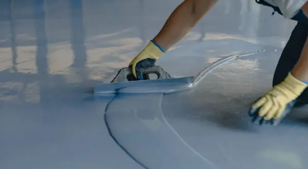 Persona con guantes amarillos aplica una capa de pintura azul sobre un suelo de terrazo con una llana.