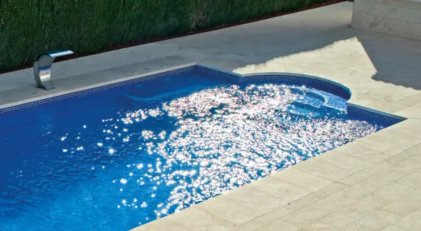 Un moderno patio con piscina de agua azul brillante, rodeado de baldosas de piedra y un seto verde. El sol crea destellos deslumbrantes en la superficie del agua.