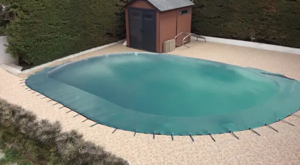 Cubierta verde protege la piscina para el invierno. Un cobertizo de madera y setos rodean el área.