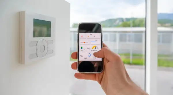 Control de domótica en casa con el móvil. Se ve un termostato en la pared y una mano manejando una app de control de clima y humedad.