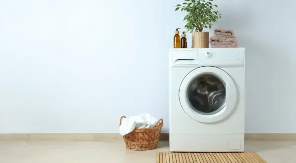Lavadora blanca con planta y toallas encima. Cerca, una cesta con ropa blanca. La altura lavadora es perfecta para el espacio.