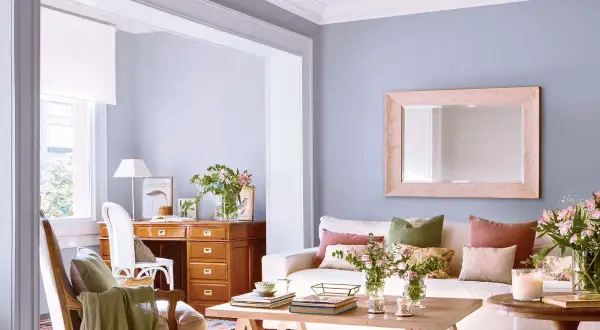 Ideas para decorar casa con poco dinero: sala acogedora con escritorio de madera, sofá blanco, cojines coloridos y flores frescas.