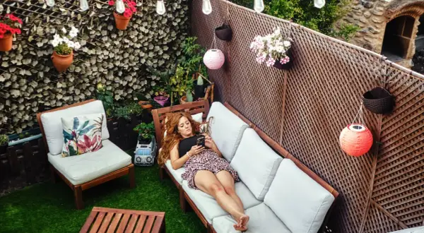 Mujer en sofá de patio, rodeada de plantas y luces. Una idea para decorar pared patio exterior con macetas colgantes y farolillos.