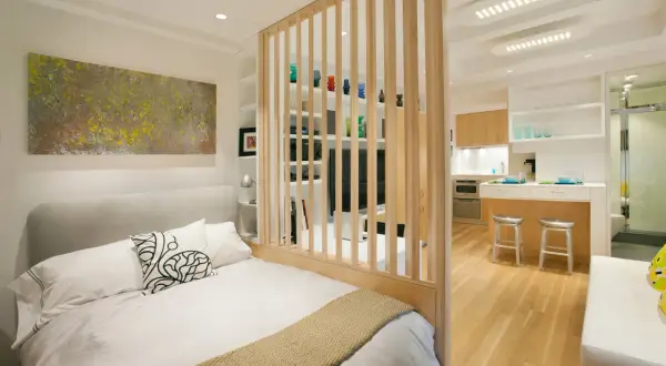 Un dormitorio moderno y funcional, parte de un diseño de interiorismo para estudios, con una cama confortable y una cocina integrada separada por una celosía de madera.