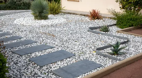 Senderos de piedra gris cruzan jardines con piedras blancas y grises, destacando plantas suculentas y un gran cactus.