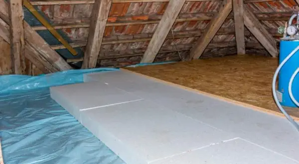 Aislamiento de suelo en una casa antigua con paneles de poliestireno sobre plástico azul. Vigas de madera y tejas visibles.