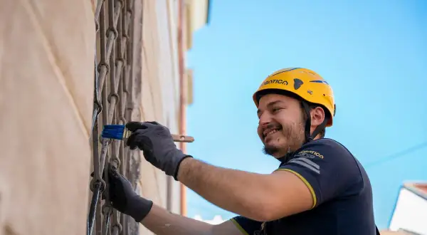 Hombre con casco amarillo sonríe mientras usa un pincel para rebozar paredes y rejas metálicas.