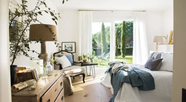 Acogedor dormitorio azul y blanco con cama, sillón y cómoda de madera. Ventanal con vistas a un jardín verde.