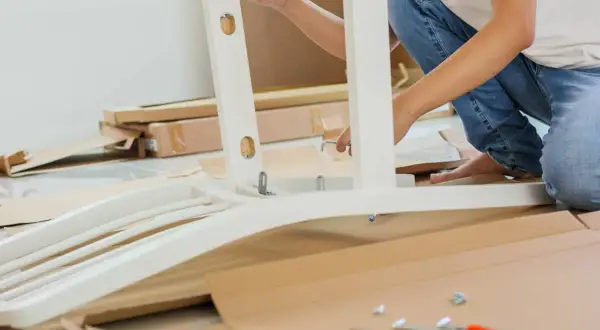 Persona ensambla mueble blanco. Se ve una pletina metálica uniendo piezas. Cajas y herramientas en el suelo.