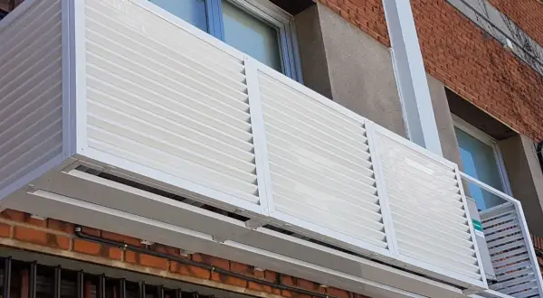 Carcasa blanca con lamas horizontales protege un aire acondicionado en fachada exterior.