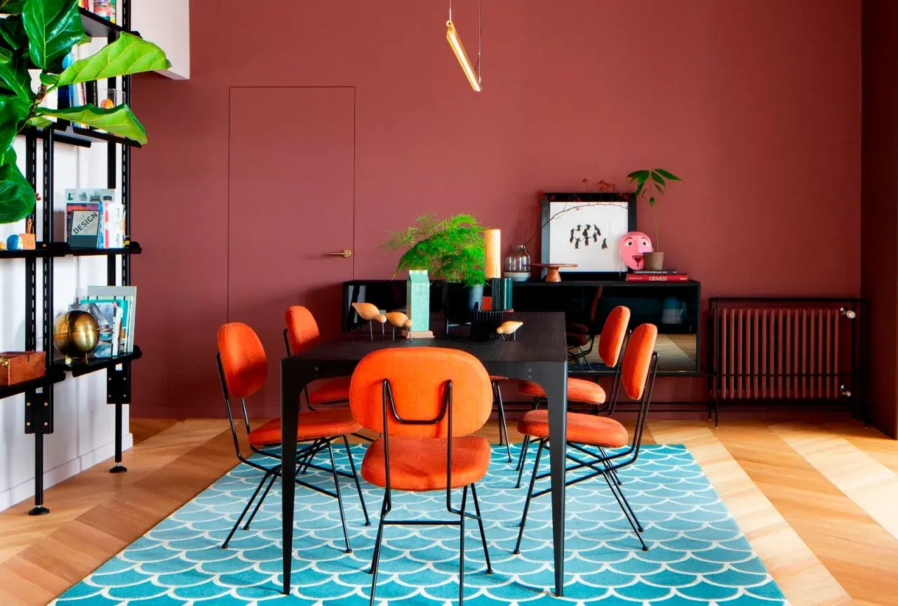 Comedor con paredes color terracota, sillas naranjas vibrantes y una alfombra azul con diseño de escamas. ¡Ideas de colores para pintar un salón audaz!