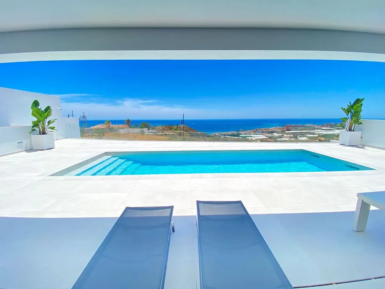 Dos tumbonas invitan a relajarse junto a una moderna piscina blanca, con el mar azul y el paisaje mediterráneo de fondo.