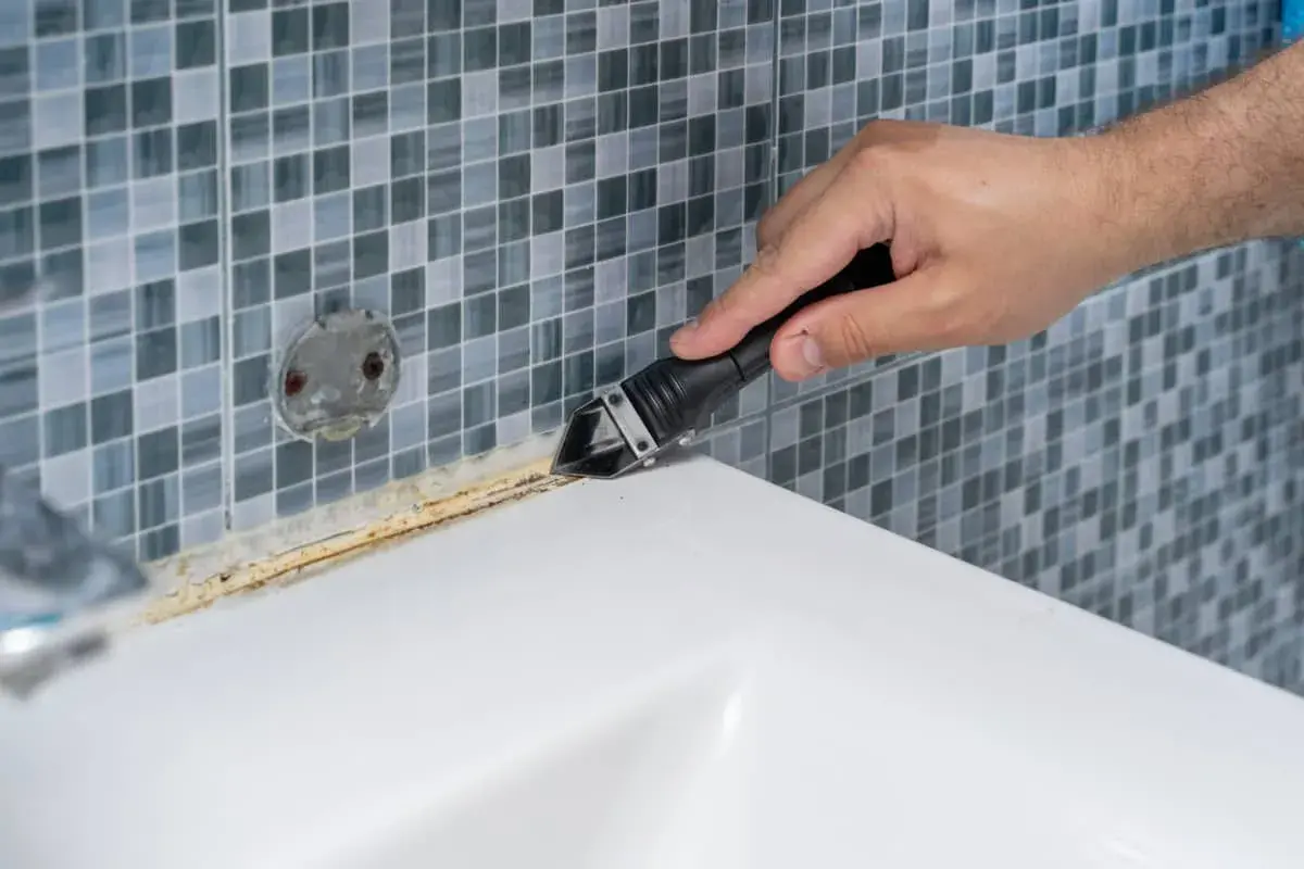 Mano usando una herramienta para quitar silicona vieja y sucia. El vinagre blanco puede ayudar a ablandar la silicona para facilitar su remoción.