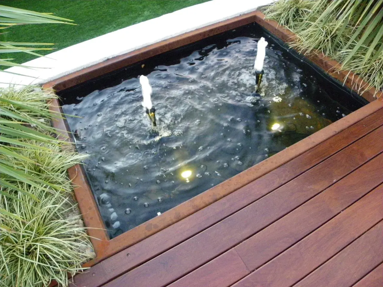 Un estanque jardín casero con dos surtidores de agua burbujeante, rodeado de vegetación y una tarima de madera.