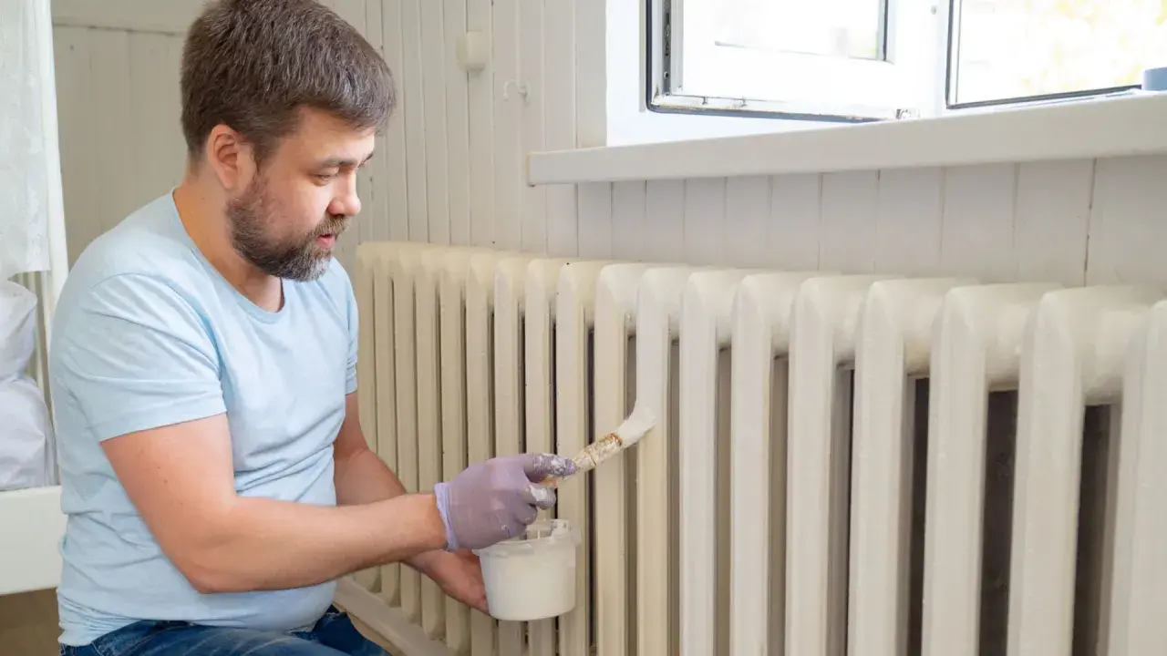 Hombre con guantes pinta radiadores de hierro con brocha y pintura blanca.