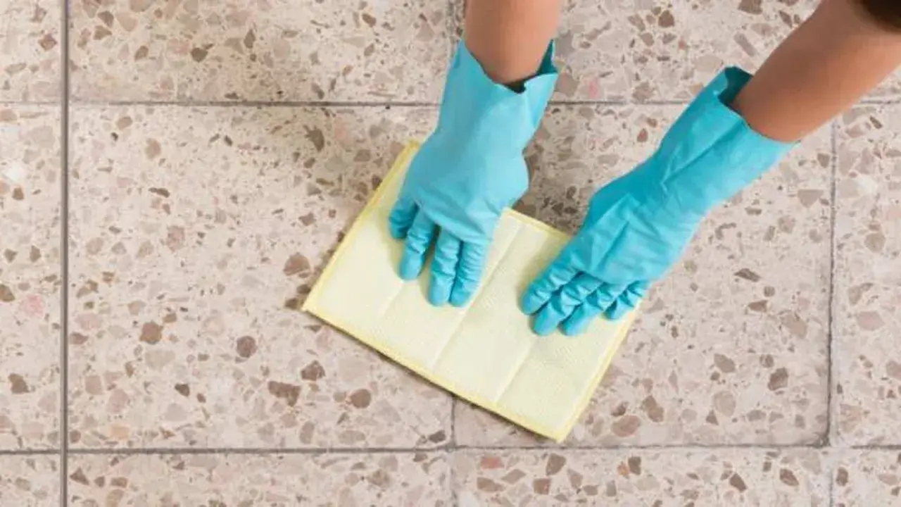 Manos con guantes azules limpiando un suelo de terrazo con un paño amarillo.