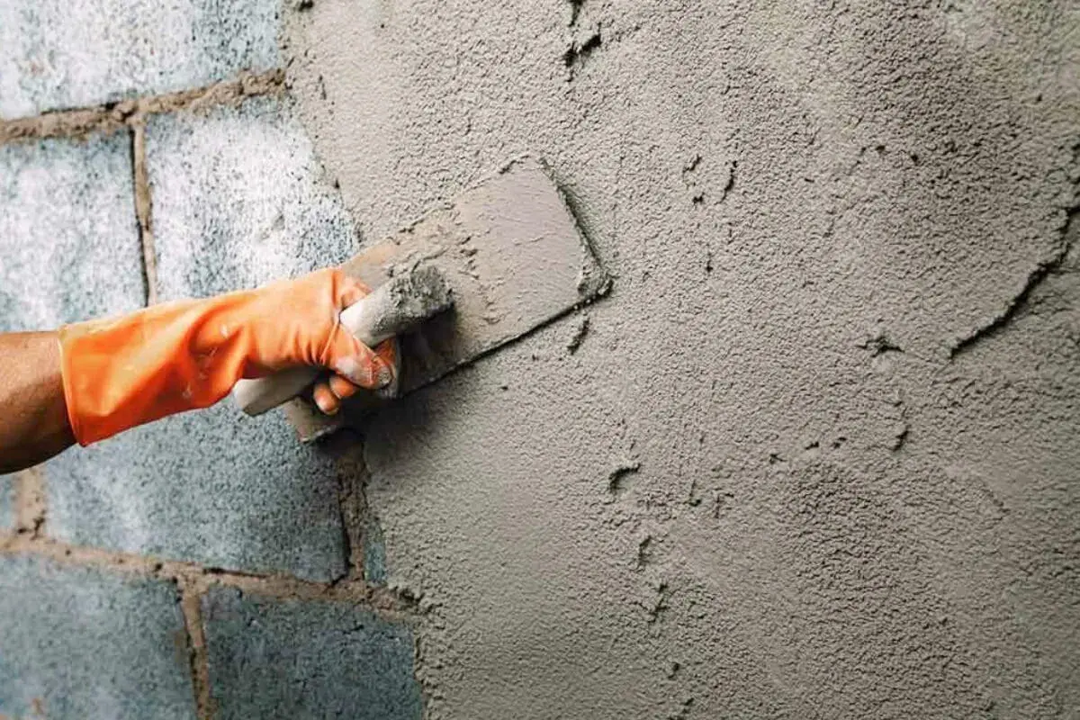 Mano con guante naranja aplica cemento para enfoscar pared exterior con llana.