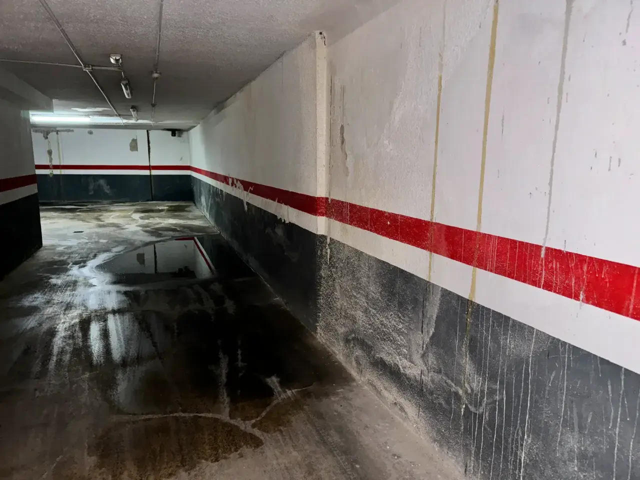 Pasillo de garaje con charcos en el suelo y marcas de humedad por filtración en las paredes pintadas con franjas rojas y grises.