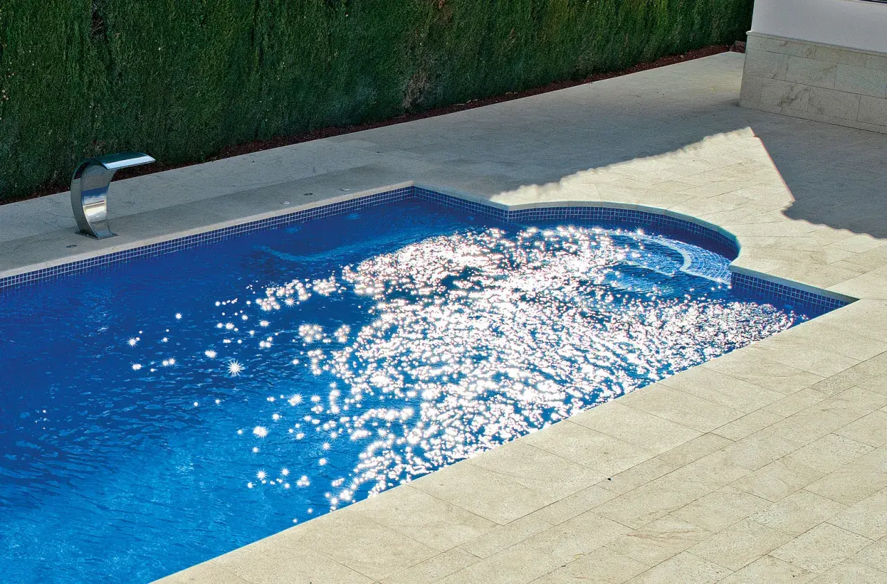 Un moderno patio con piscina de agua azul brillante, rodeado de baldosas de piedra y un seto verde. El sol crea destellos deslumbrantes en la superficie del agua.