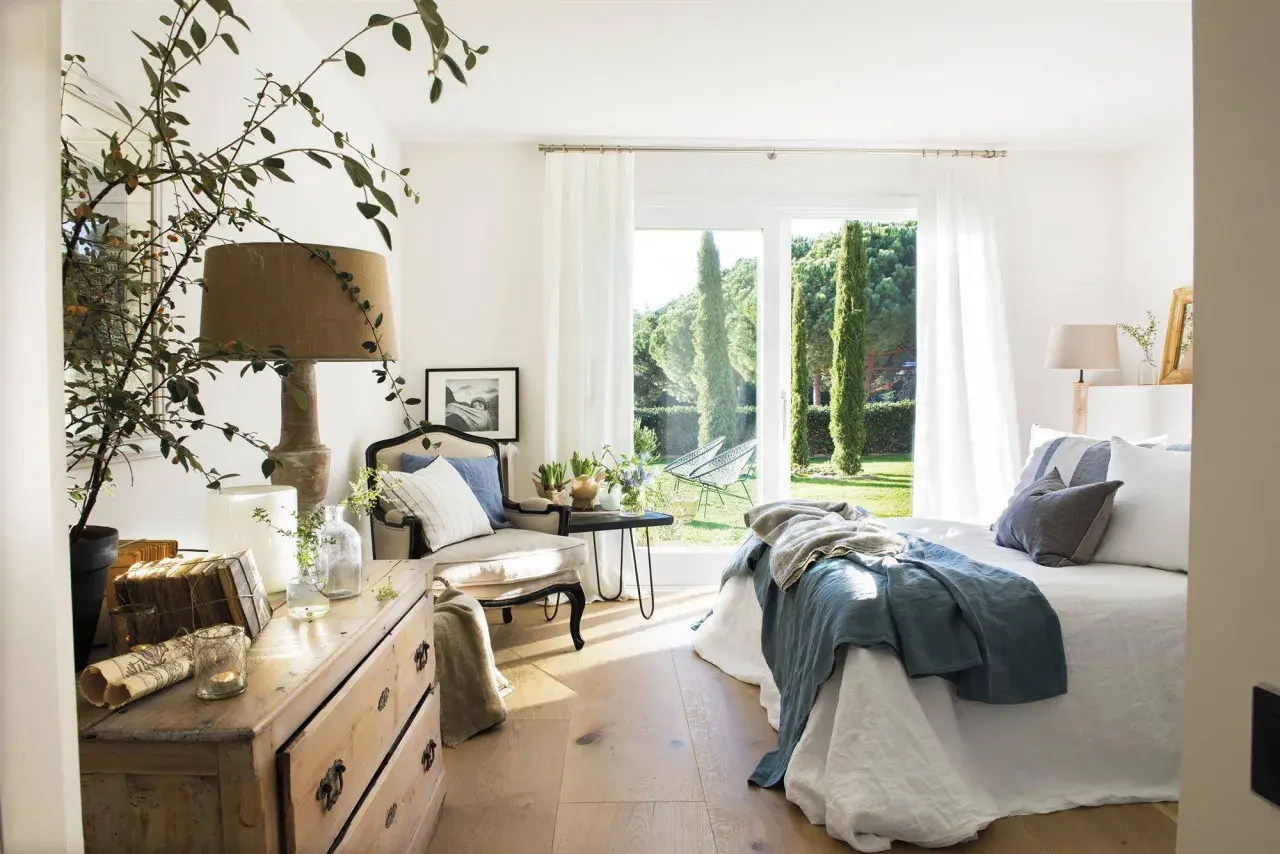 Acogedor dormitorio azul y blanco con cama, sillón y cómoda de madera. Ventanal con vistas a un jardín verde.