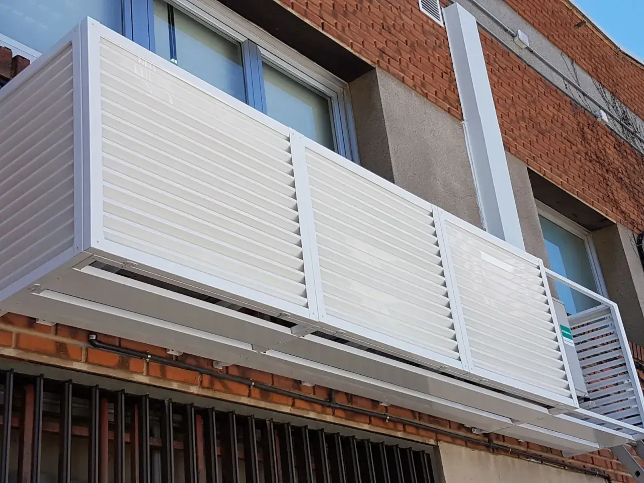 Carcasa blanca con lamas horizontales protege un aire acondicionado en fachada exterior.