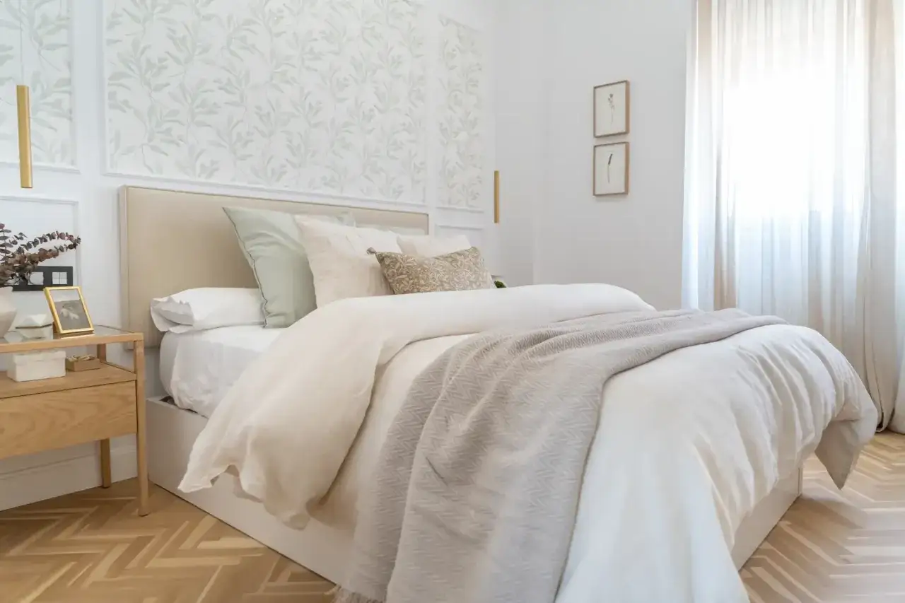 Acogedor dormitorio con papel pintado de hojas verdes en el cabecero, cama vestida con ropa de cama blanca y manta gris.