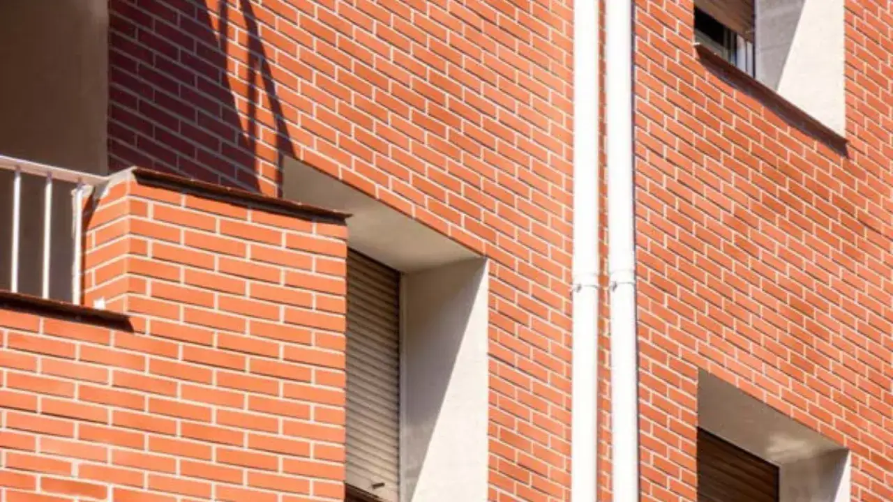 Detalle de la fachada edificio de ladrillo naranja con ventanas y tuber&iacute;as blancas.