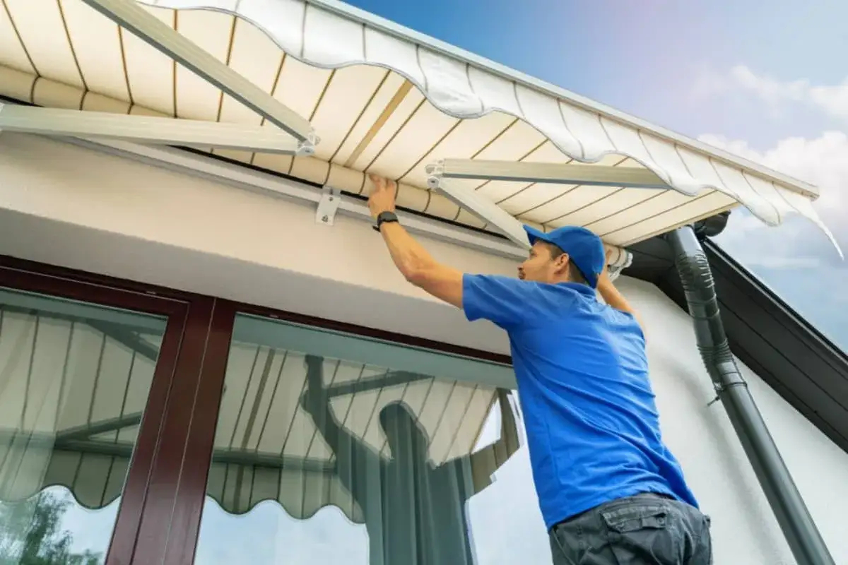 Técnico ajustando la tela de un toldo exterior. El toldo es de rayas beige y blanco, con un borde ondulado. El hombre lleva una gorra azul y una camiseta azul.
