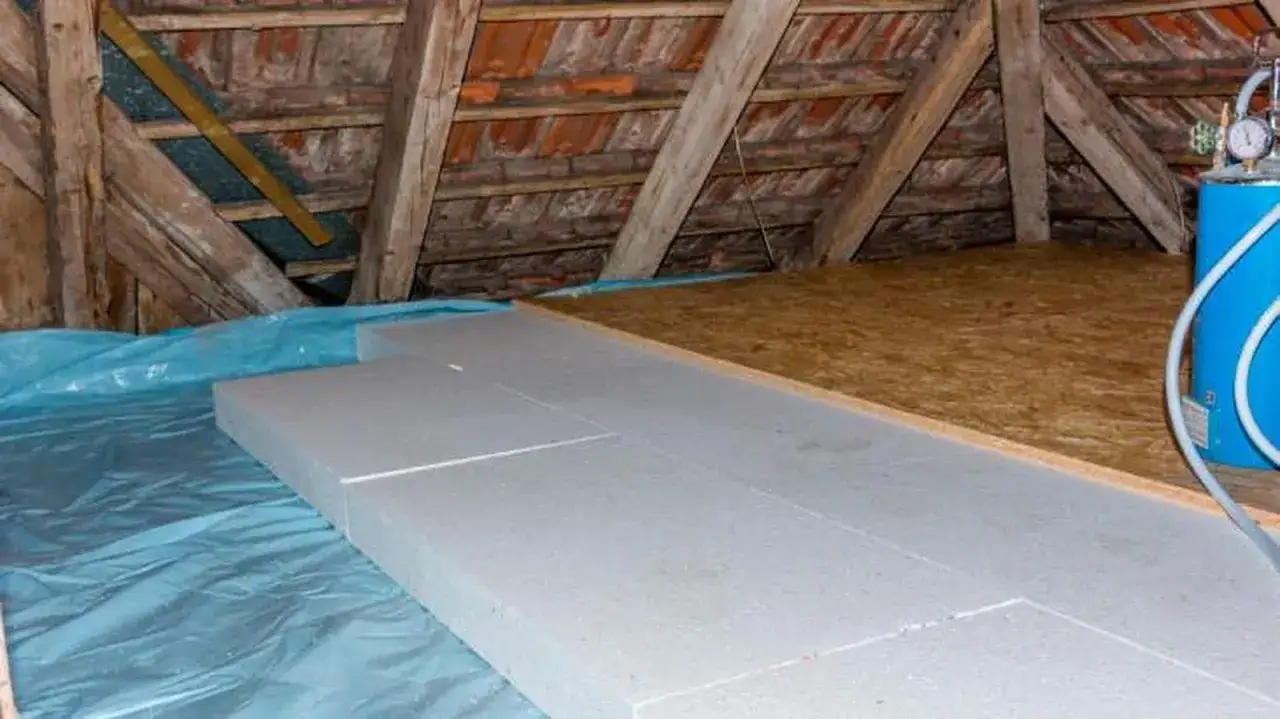 Aislamiento del suelo de una casa antigua con poliestireno expandido sobre lámina azul. Vigas de madera y tejas visibles.
