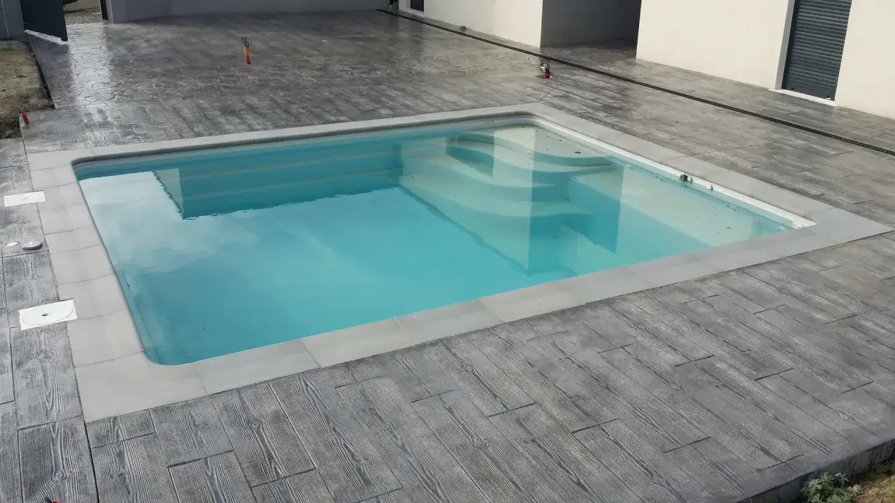 Piscina de agua cristalina con escalones integrados, rodeada de un suelo de hormig&oacute;n impreso que imita madera. Lista para hacer piscina y disfrutar del verano.