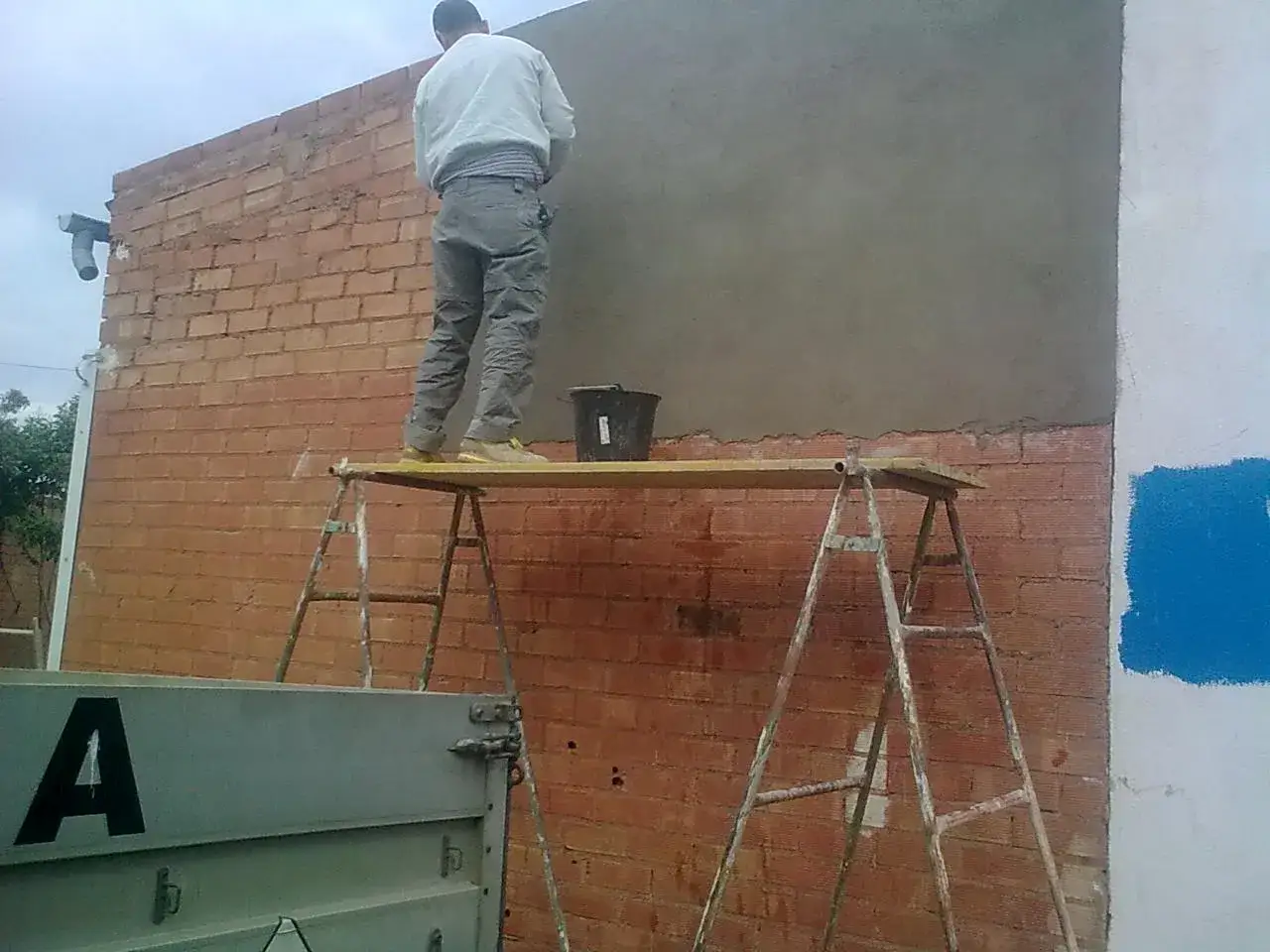 Obrero en andamio enfoscando pared exterior de ladrillo. Se ve un cubo negro y parte de un contenedor.