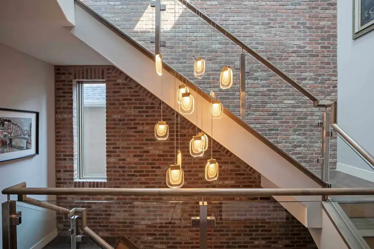 Moderna cascada de luces colgantes para decorar subida de escalera interior. Ladrillo visto y barandillas de madera y cristal.