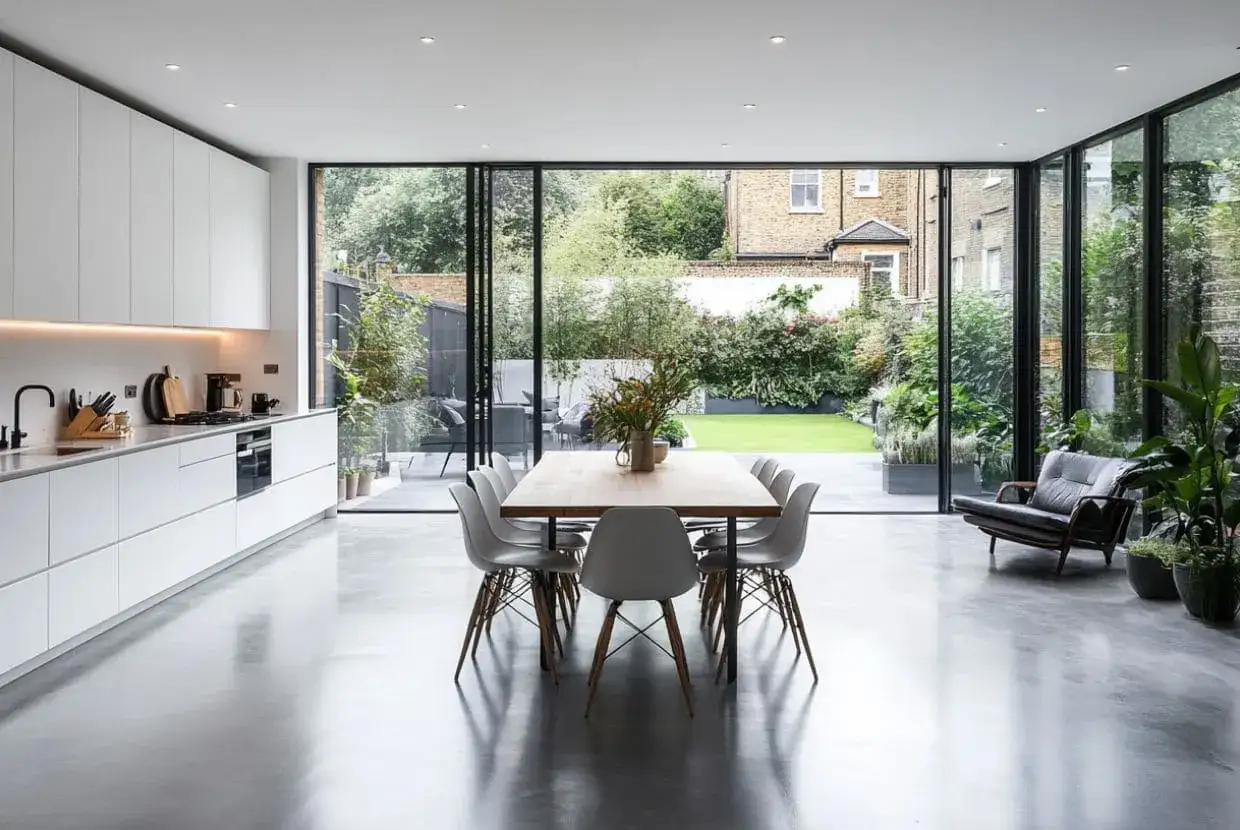 Cocina moderna con encimeras blancas, mesa de comedor y sillas, y un sill&oacute;n de cuero, todo sobre un suelo de cemento pulido que refleja la luz del jard&iacute;n.