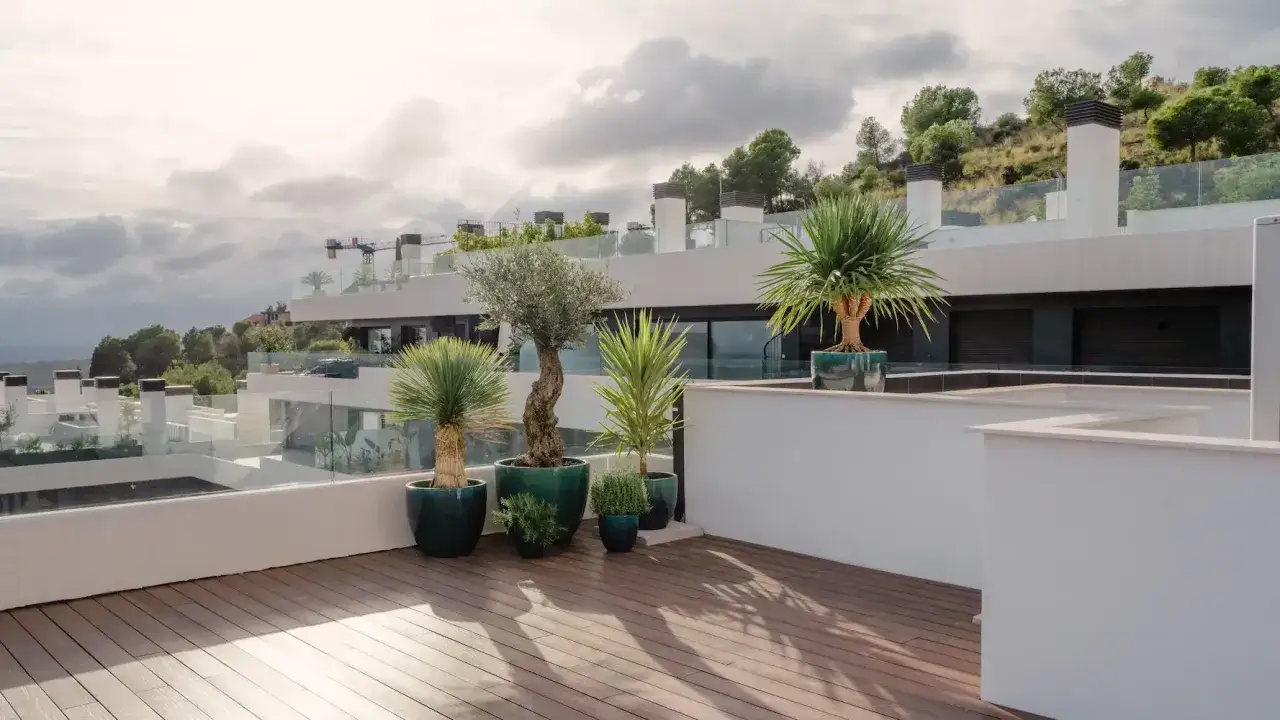 Un moderno jardín urbano en una terraza con árboles en macetas, olivo y palmeras, bajo un cielo nublado.