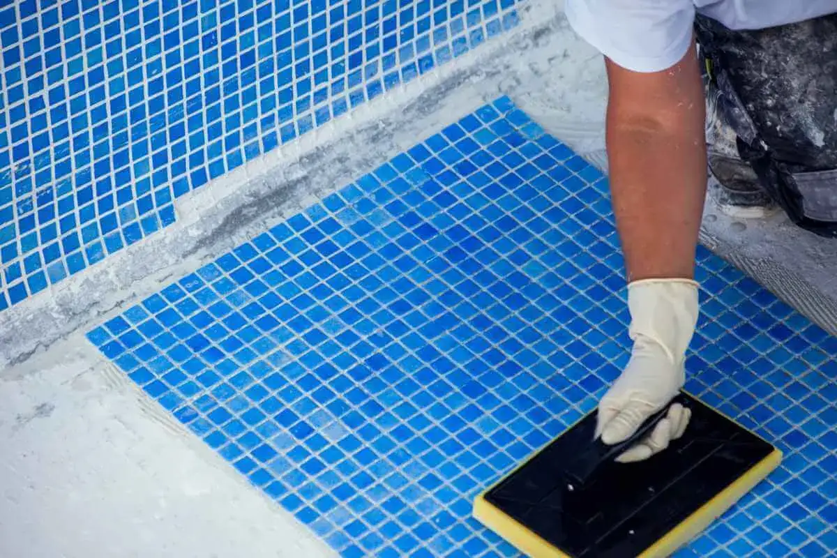 Un trabajador con guantes aplica lechada a las baldosas azules de una piscina, un paso clave para arreglar piscina y dejarla lista para el verano.