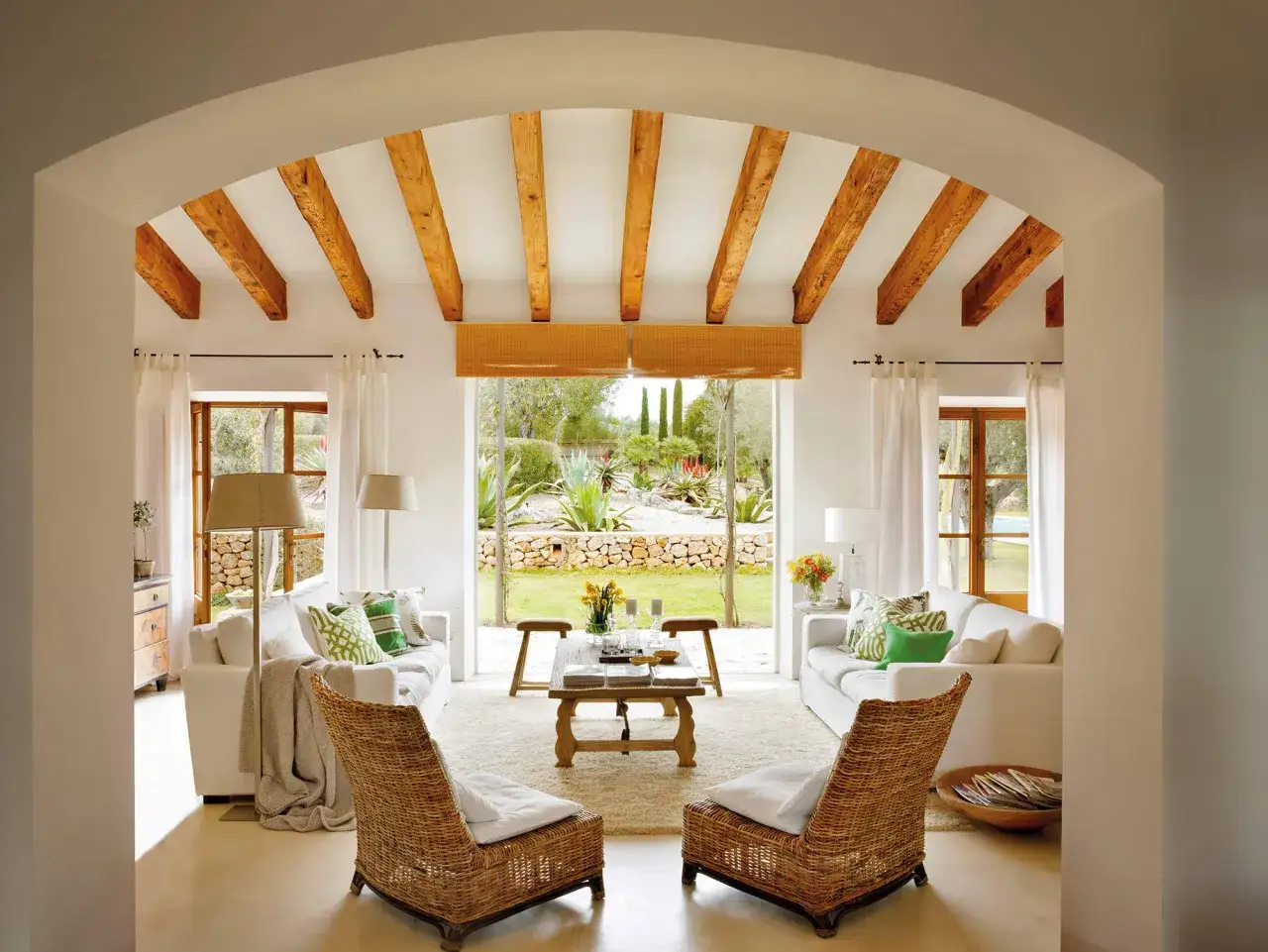 Sal&oacute;n con decoraci&oacute;n ibicenca, vigas de madera, sof&aacute;s blancos y sillones de mimbre, con vistas a un jard&iacute;n mediterr&aacute;neo.