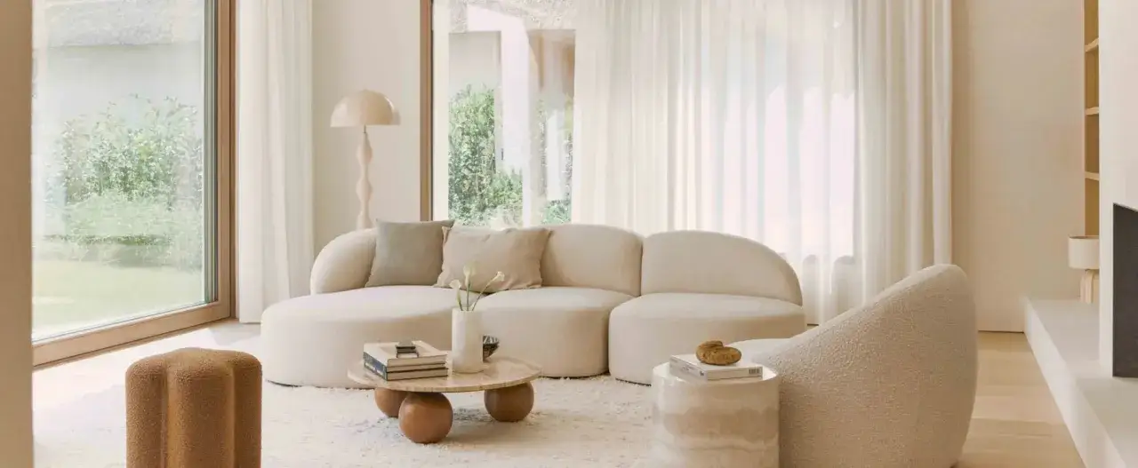 Un sofá curvo y acogedor con cojines neutros. Una mesa de centro baja con libros y un jarrón. Un puf marrón con forma de flor. Ideas de que poner detras del sofa para un salón moderno.