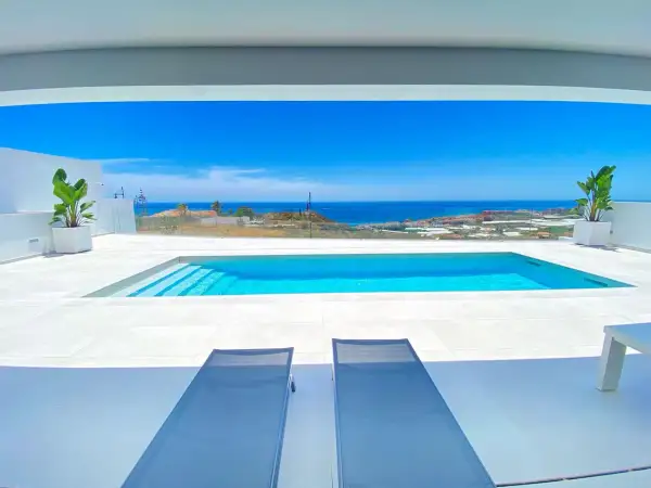 Dos tumbonas invitan a relajarse junto a una moderna piscina blanca, con el mar azul y el paisaje mediterráneo de fondo.