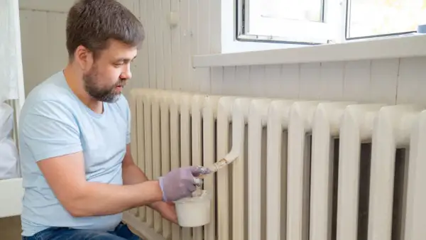Hombre con guantes pinta radiadores de hierro con brocha y pintura blanca.