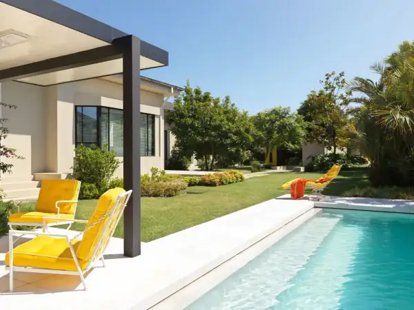 Un moderno jardín con piscina y porche, donde dos sillas amarillas invitan a relajarse bajo el sol.