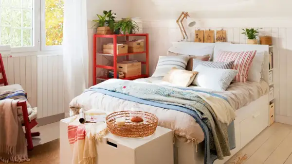 Acogedora habitación pequeña para adultos con cama, estantería roja y ventana luminosa.