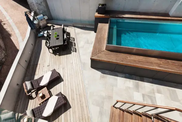 Terraza con tumbonas, mesa y barbacoa junto a una piscina elevada de obra con agua azul.