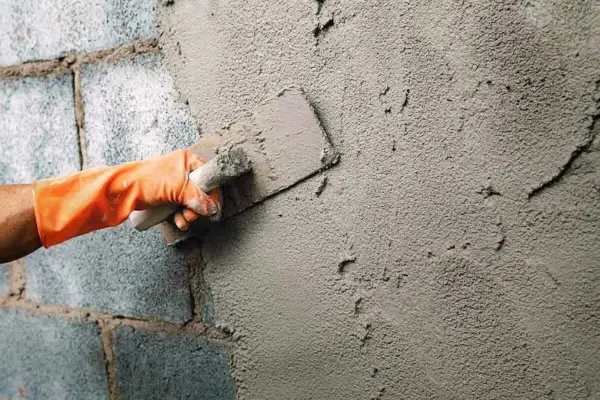 Mano con guante naranja aplica cemento para enfoscar pared exterior con llana.