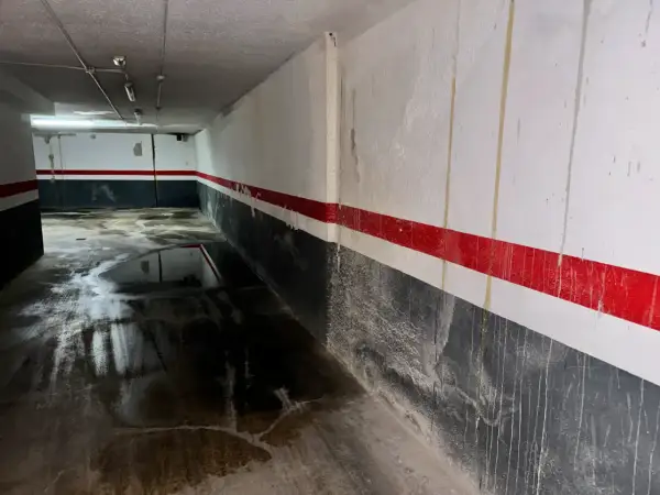 Pasillo de garaje con charcos en el suelo y marcas de humedad por filtración en las paredes pintadas con franjas rojas y grises.