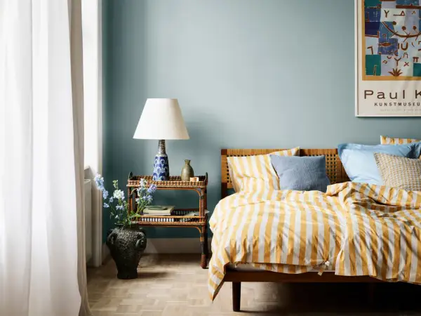 Habitación con paredes azul claro, cama con ropa de cama a rayas amarillas y blancas, y mesita de noche de ratán. Ideas de colores para habitaciones.