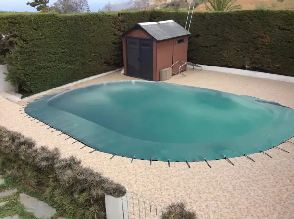 Cubierta verde protege la piscina para el invierno. Un cobertizo de madera y setos rodean el área.