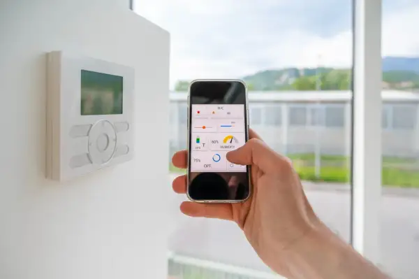 Control de domótica en casa con el móvil. Se ve un termostato en la pared y una mano manejando una app de control de clima y humedad.