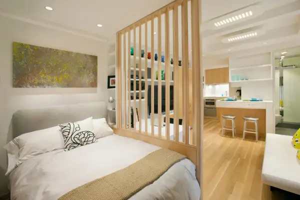 Un dormitorio moderno y funcional, parte de un diseño de interiorismo para estudios, con una cama confortable y una cocina integrada separada por una celosía de madera.